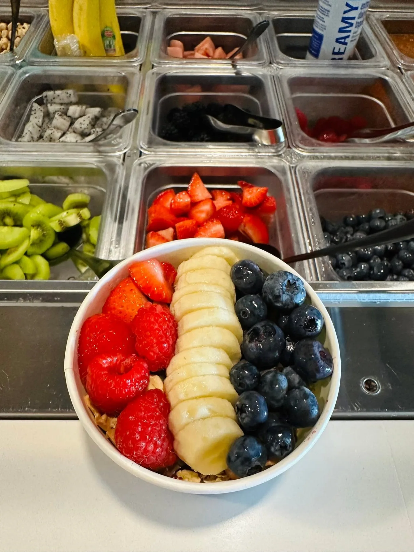 Açaí bowl with raspberries and strawberries on one side, banana slices lined up down the middle and blueberries on the other, at the AMI FroYo toppings bar