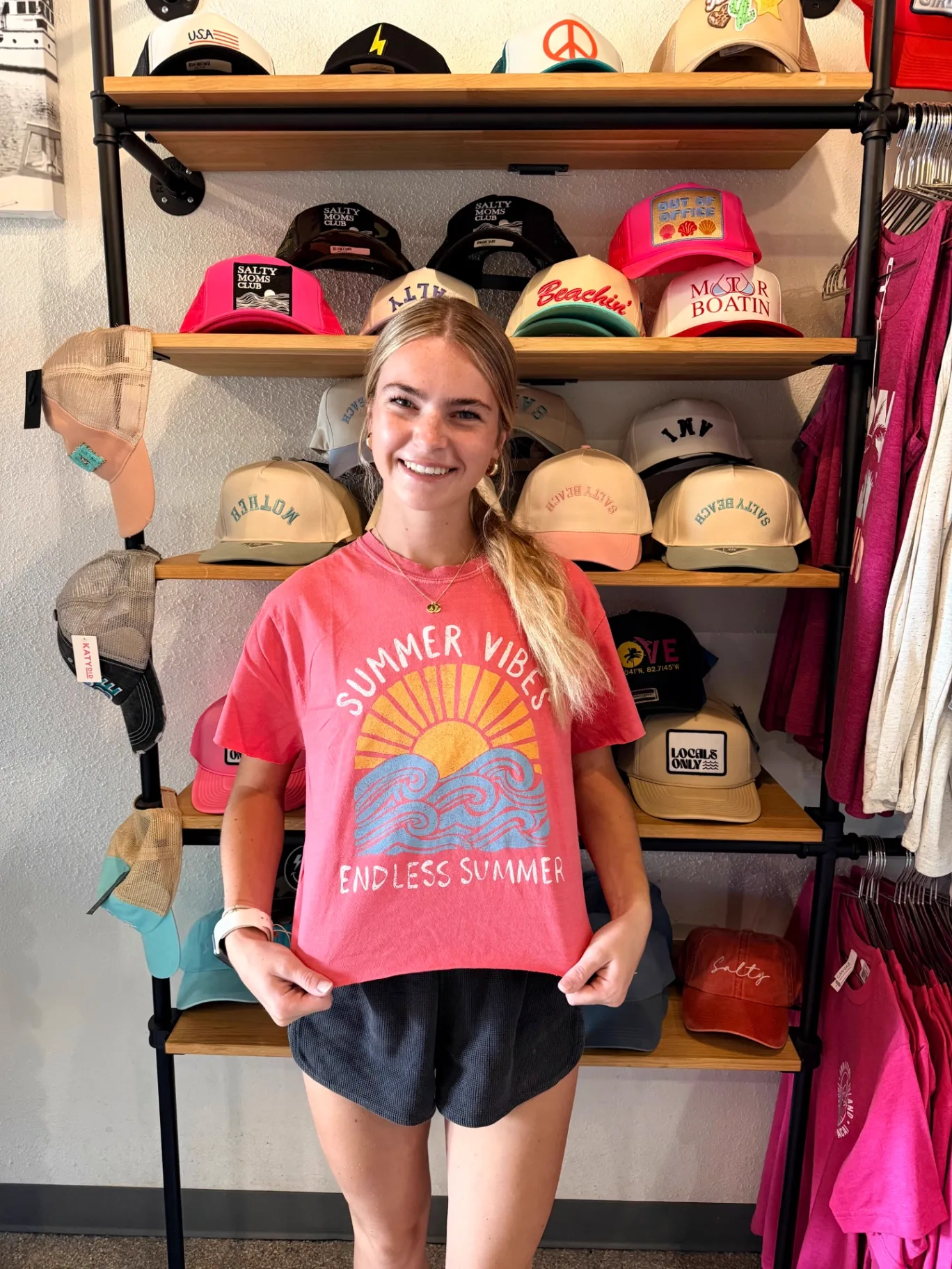 Team member showing the pink Summer Vibes tee in front of the hat wall