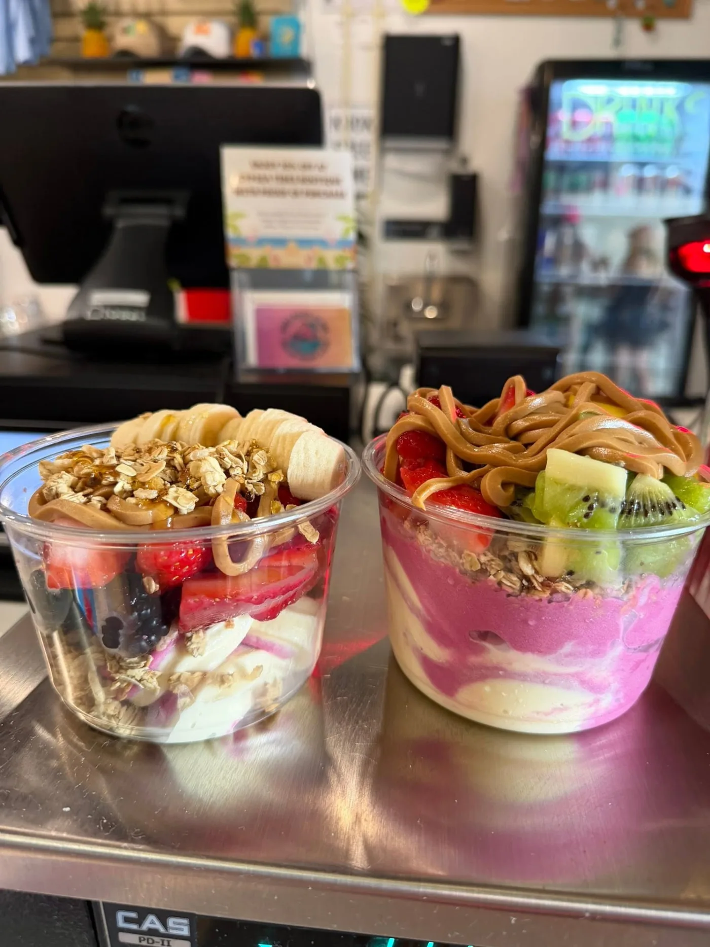 Two loaded bowls with peanut butter drizzle, strawberries, kiwi, granola and banana on the counter